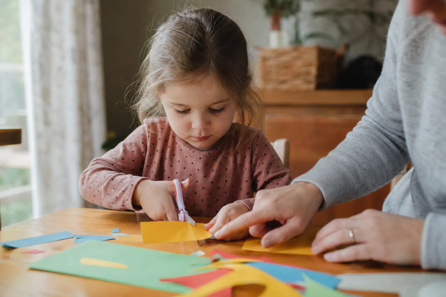Un jeune enfant concentré découpe une bande de papier coloré avec de petits ciseaux, aidé par un parent.
