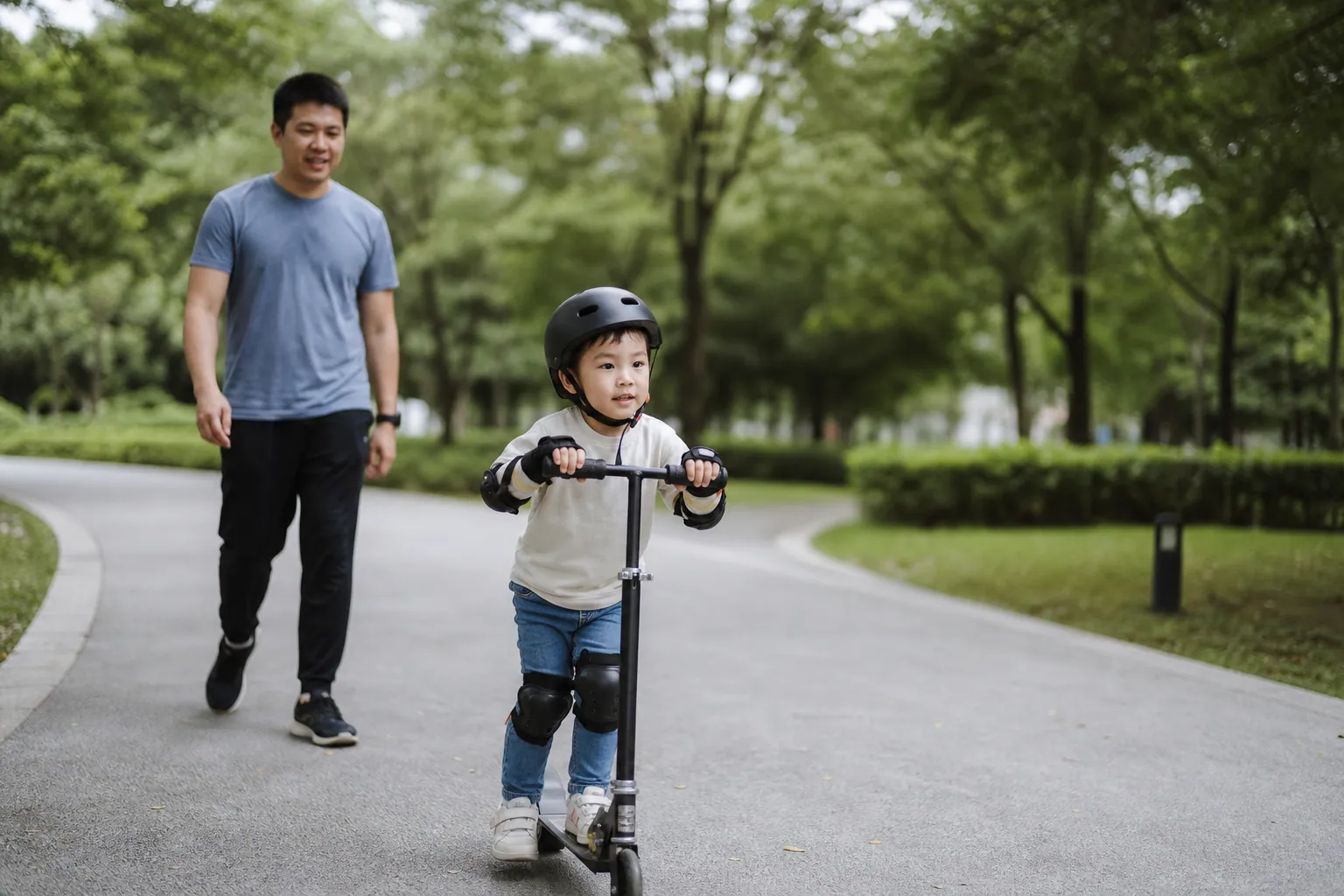 Un enfant casqué roule en trottinette sur une allée, un parent le suit à quelques pas.