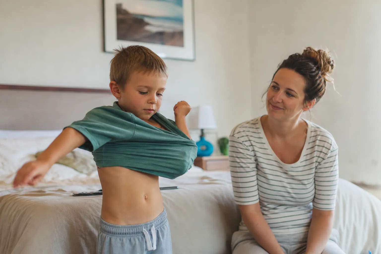 Un enfant enfile seul un tee-shirt dans sa chambre sous le regard encourageant d'un parent.