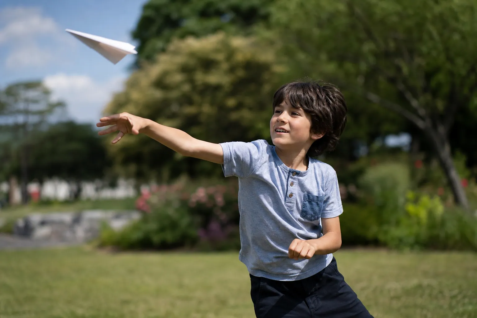 Un enfant lance un avion en papier blanc dans un parc ensoleillé.