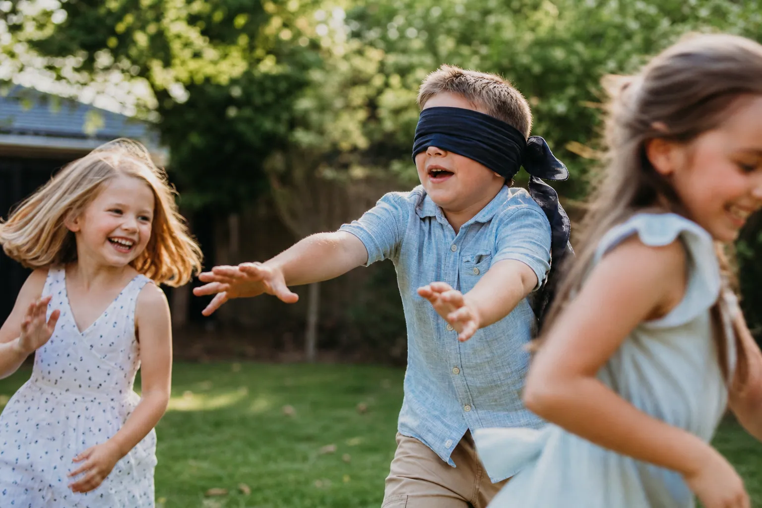 Un enfant les yeux bandés tente d'attraper d'autres enfants qui l'évitent dans un jardin.