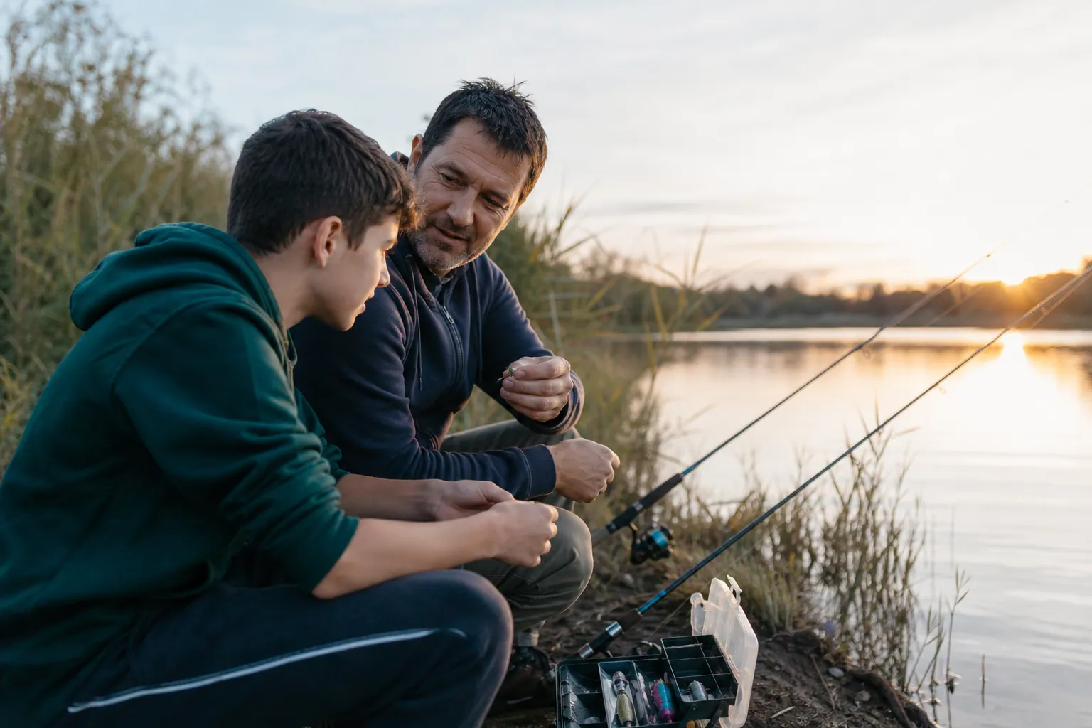 Un parent et son adolescent pêchent au bord d’un lac avec leur matériel, au lever du jour.