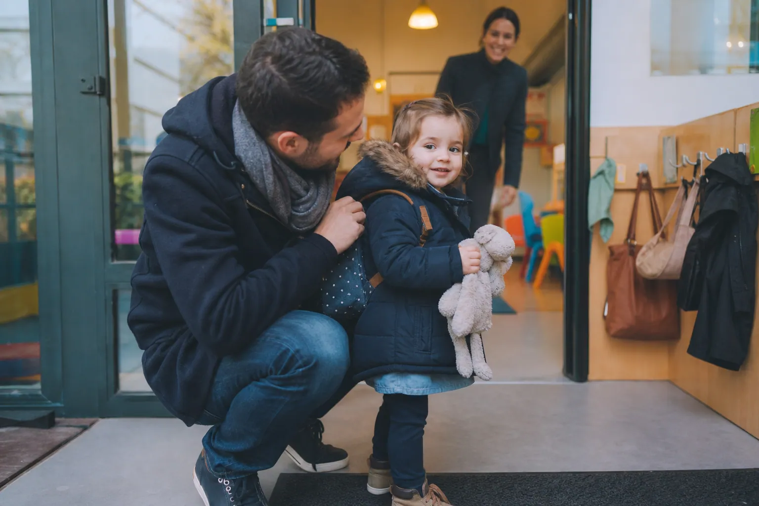 Parent rassurant son tout-petit à l’entrée de la crèche, avec une professionnelle à l’accueil.