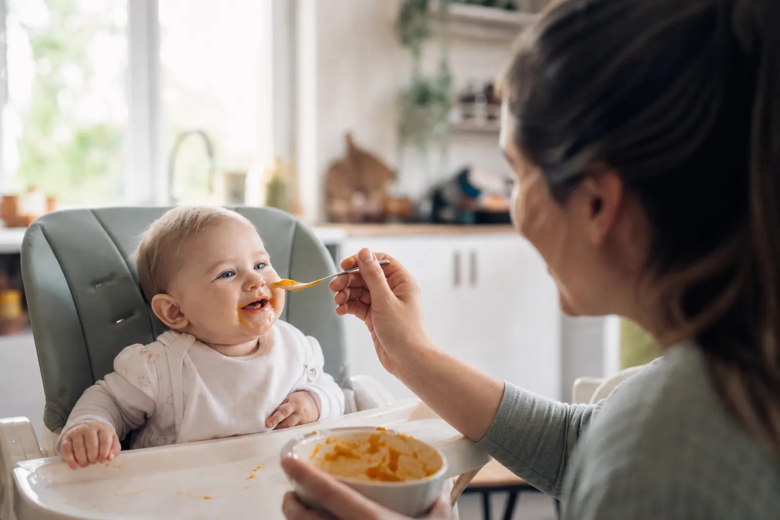 Bébé en chaise haute goûtant une purée de légumes donnée par un parent dans une cuisine lumineuse.