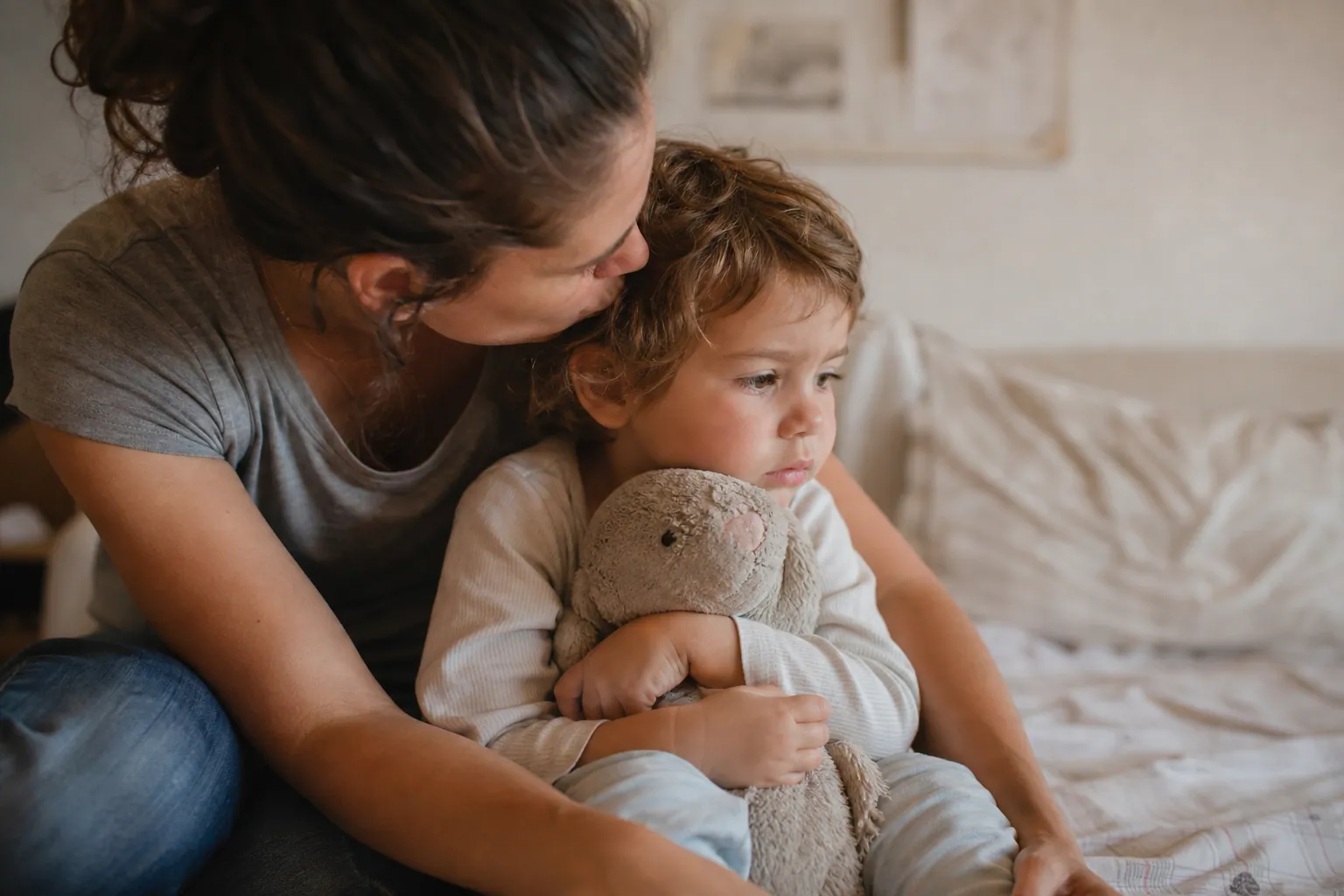 Parent réconfortant un enfant qui serre son doudou contre lui dans une chambre baignée de lumière chaude.