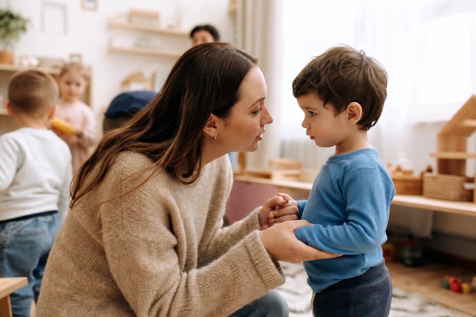 Éducatrice de crèche agenouillée, parlant calmement à un jeune enfant dans une salle de jeux.