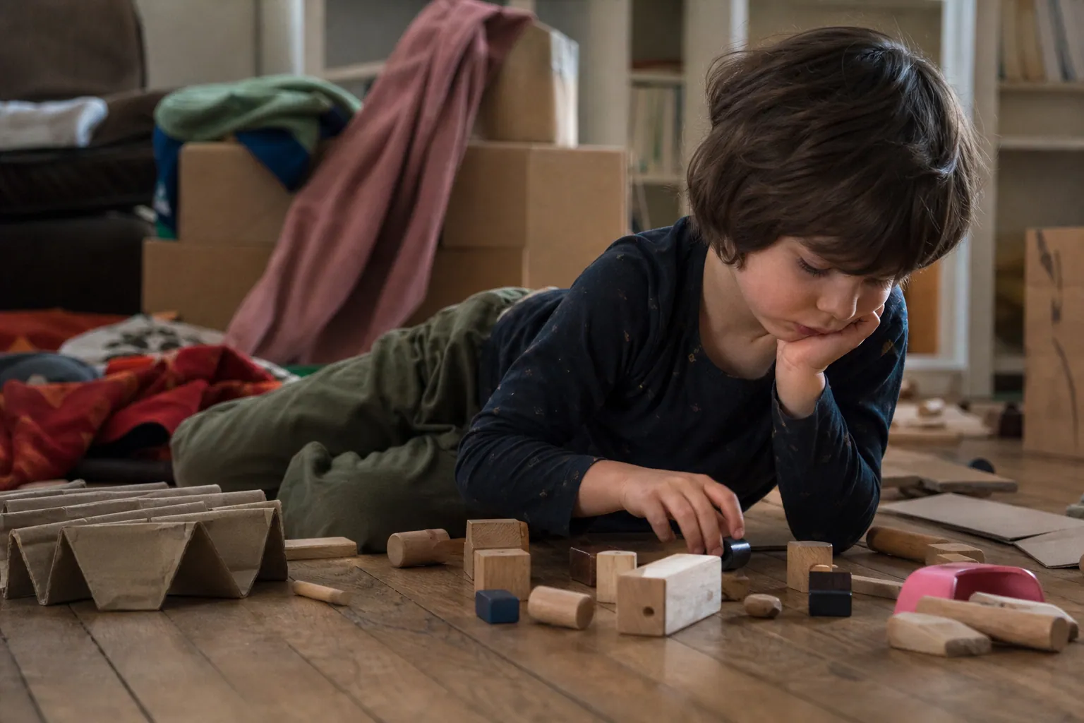 Enfant allongé au sol jouant avec des objets simples, cartons et tissus, sans écran, lumière du jour.