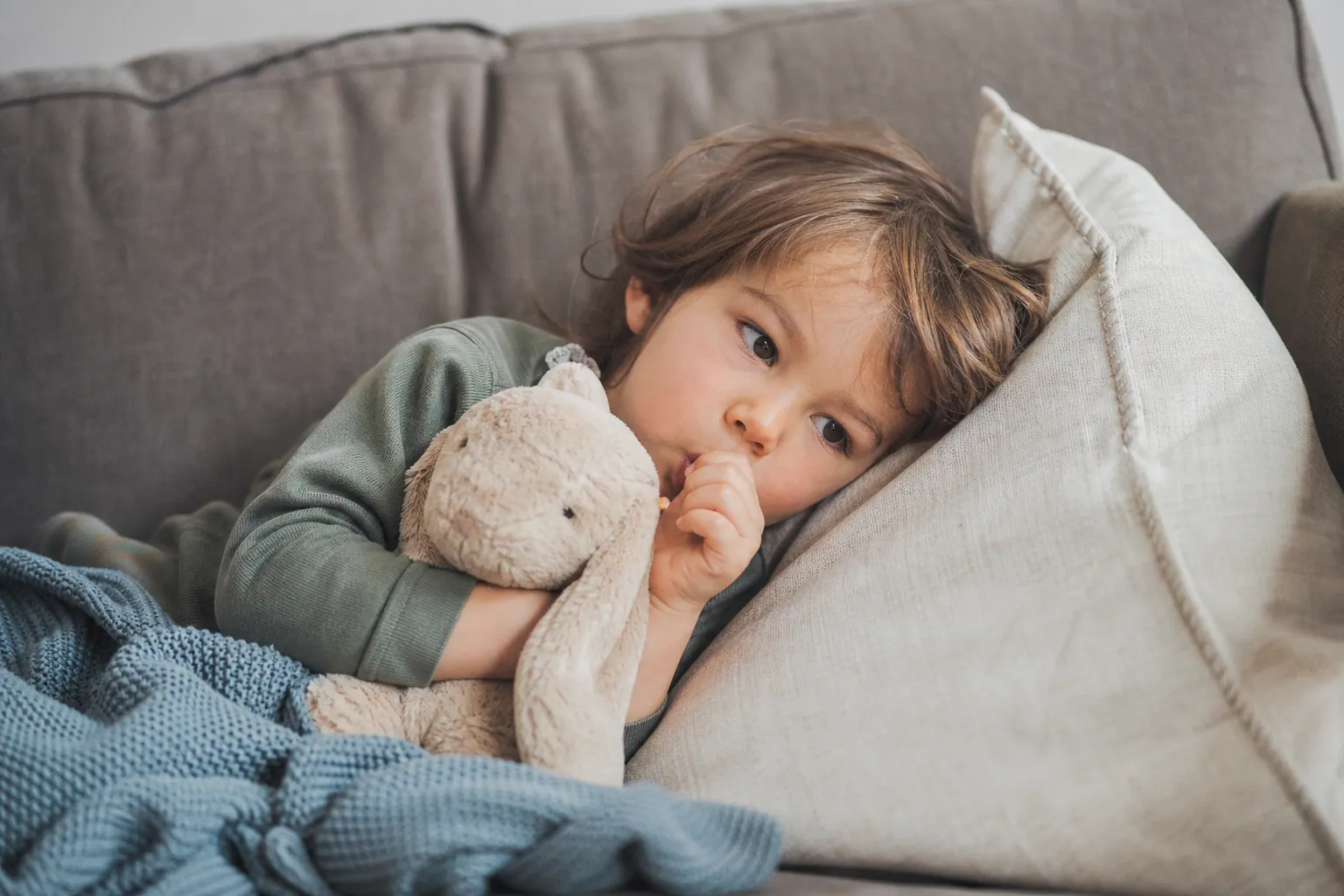 Jeune enfant suçant son pouce en câlinant une peluche, dans la lumière douce d'un salon familial.
