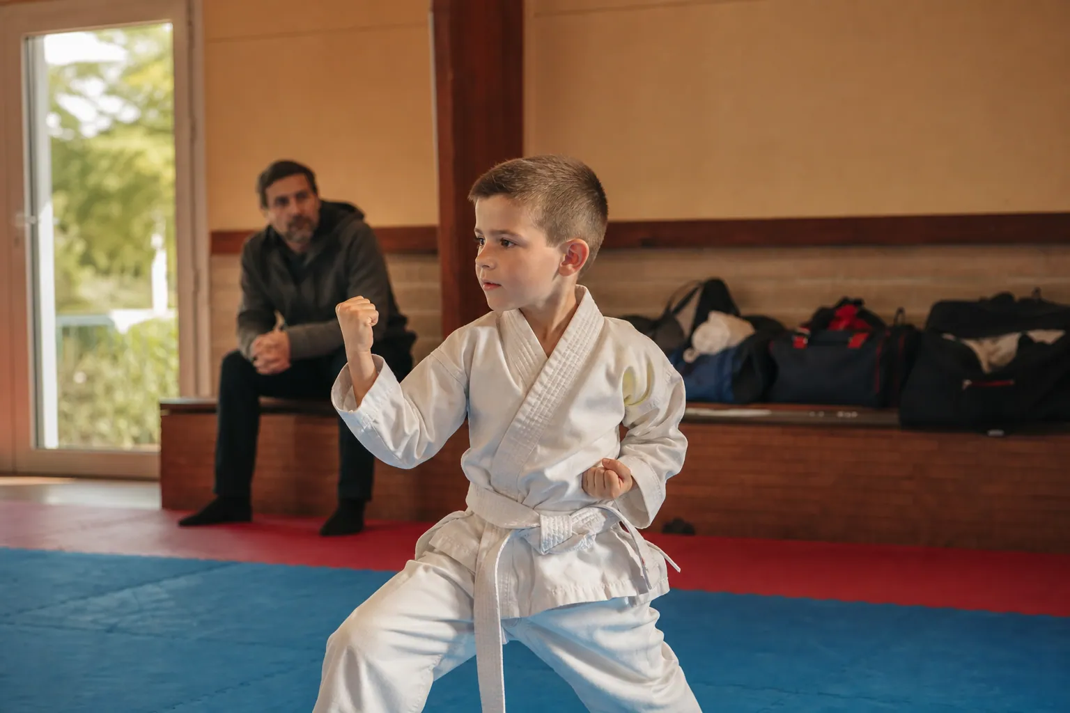 Enfant en kimono de karaté concentré dans un dojo, avec un parent en arrière-plan.