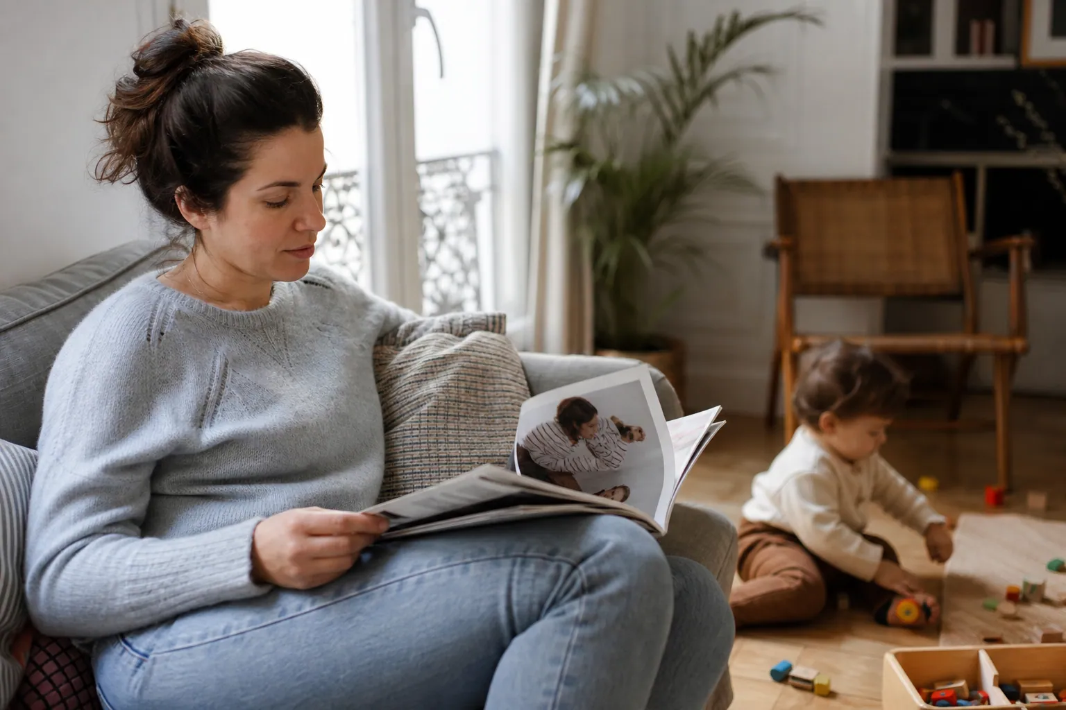 Parent feuilletant un magazine parental dans un salon familial chaleureux, avec un enfant jouant à côté.