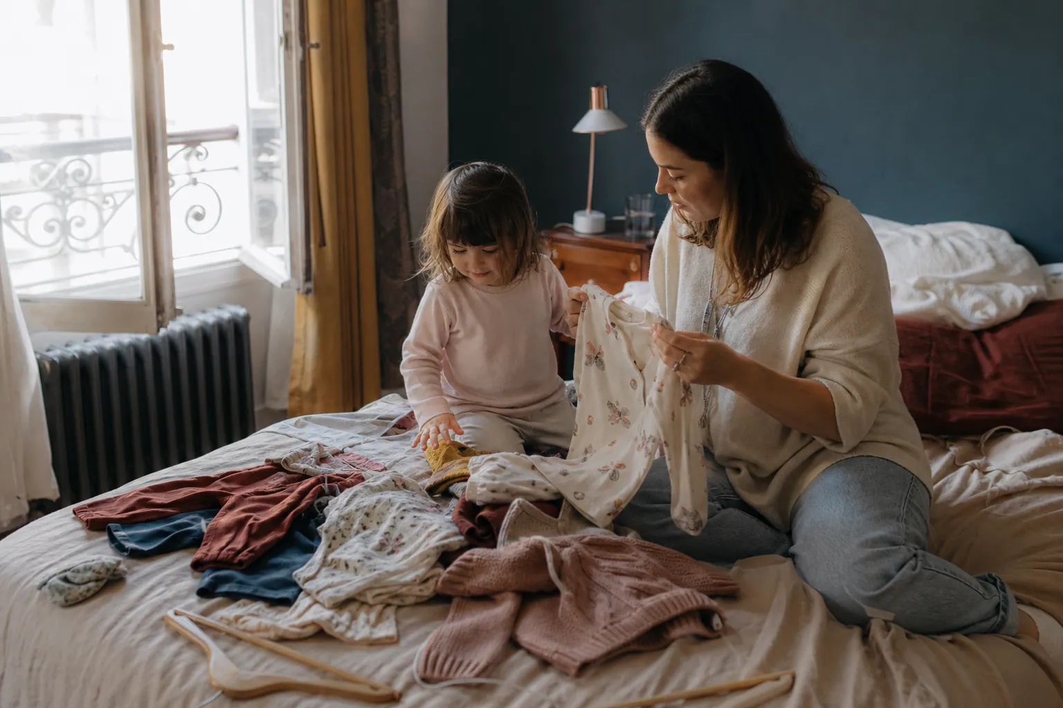 Parent et enfant choisissant des vêtements colorés sur un lit dans une chambre lumineuse.