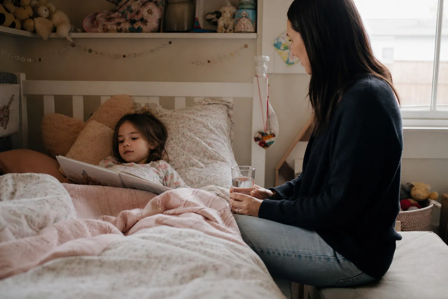 Un enfant se repose dans son lit avec un livre, tandis qu'un parent assis à côté lui tend un verre d'eau.